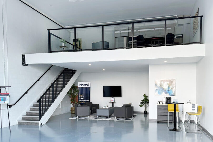 A spacious, modern commercial space with a high ceiling and polished light coloured floor. The area includes a mezzanine level with glass railings, accessible by a staircase on the left side. Below the mezzanine, there is a seating area with several grey armchairs arranged around a low coffee table. A flat-screen TV is mounted on the wall, with some plants and artwork in the same area.