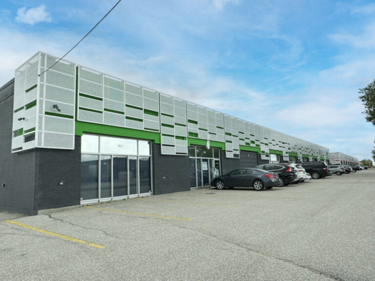 A row of commercial condos with cars in front of them, on a partly cloudy day. The building exterior features a grey and white colour scheme with bright green accents. The condos are situated on a paved lot.