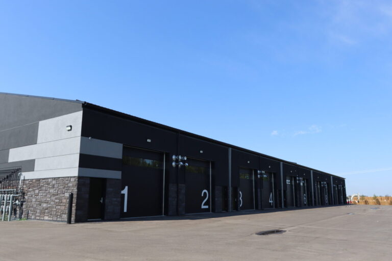 A row of modern commercial condos with large numbered doors, ranging from 1 to 8, on a clear day. The building exterior features a sleek, dark color scheme with contrasting light grey accents and stonework along the base. The garage is situated on a wide, paved lot under a bright blue sky.