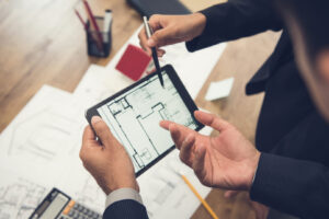 Property manager. Two business professionals reviewing building floor plans on a tablet, with architectural blueprints and office supplies spread out on a desk in the background.